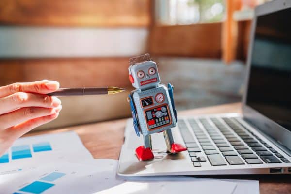 Un jouet robot rétro se tient sur le clavier de l’ordinateur portable tandis que la main tient le stylo, symbolisant la créativité et la technologie dans l’espace de travail moderne et les agents IA autonomes en 2026. Crédits iStock : jabkitticha Référence de la photo : 2240460210